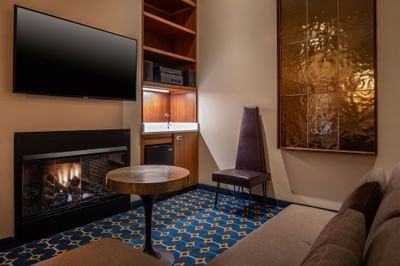 TV mounted above a fireplace beside a wooden shelf unit & a round table with a chair in the Suite at Harborside Inn Boston