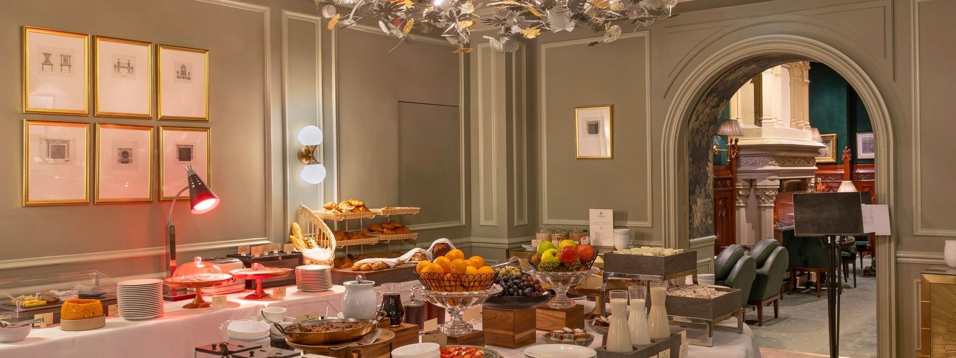 Juices and cereal by stacked white plates under a glowing leaf chandelier in the buffet at Warwick Hotels & Resorts
