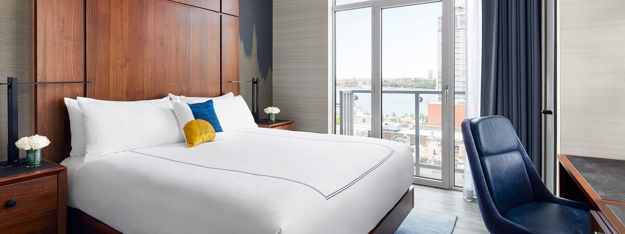 Modern hotel room with a large bed, blue chair, and balcony overlooking a cityscape.