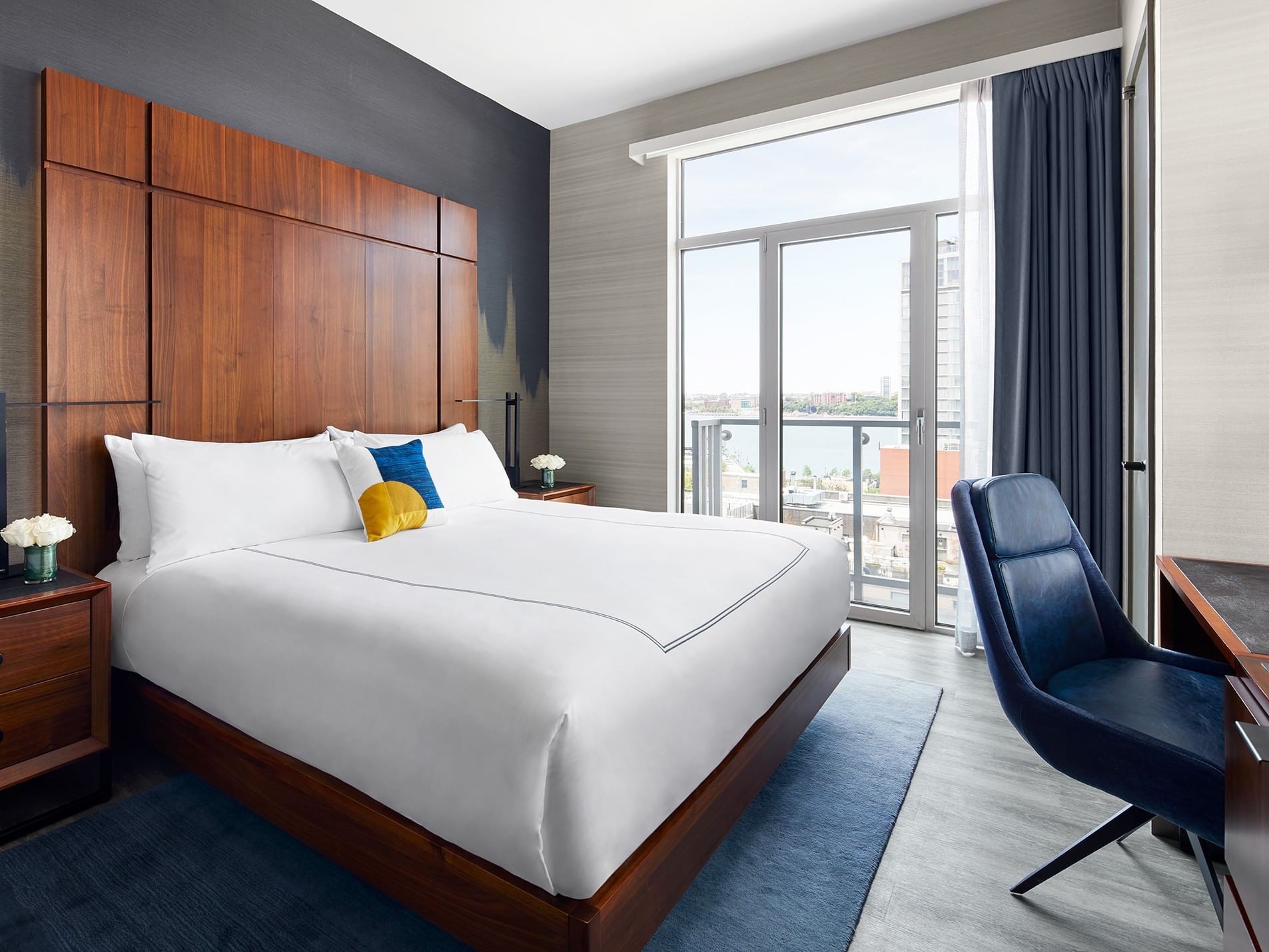 Modern hotel room with a large bed, blue chair, and balcony overlooking a cityscape.