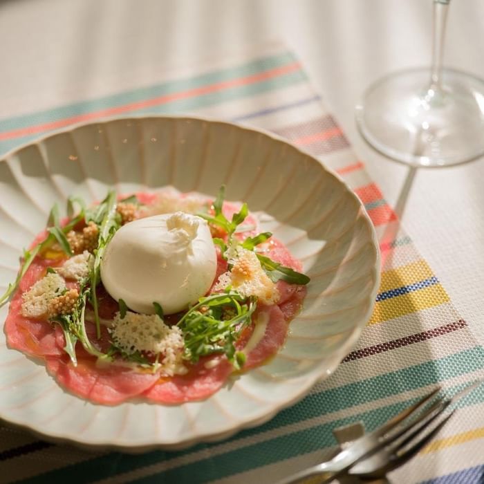 Piatto di prosciutto crudo con mozzarella, rucola e pane grattugiato, con forchetta e calice di vino.