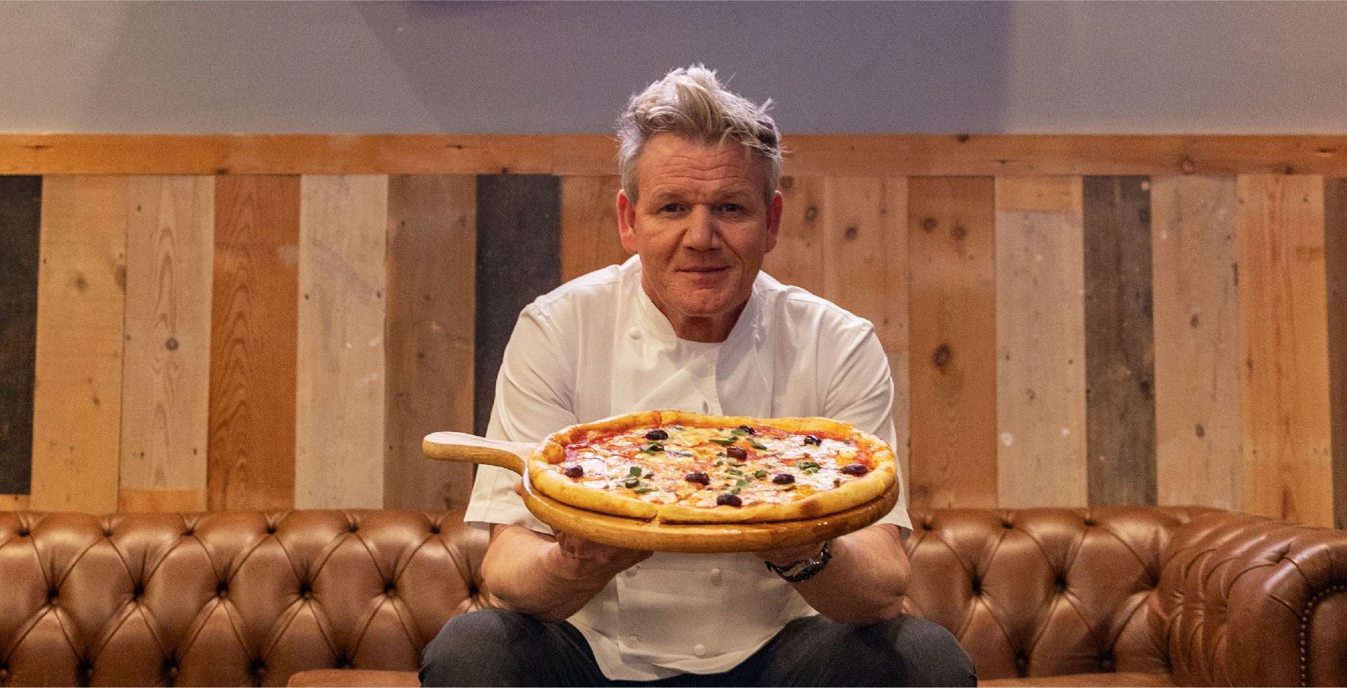 Chef Gordon Ramsay smiling and presenting a hot pizza topped with olives and basil at Sunway Resort