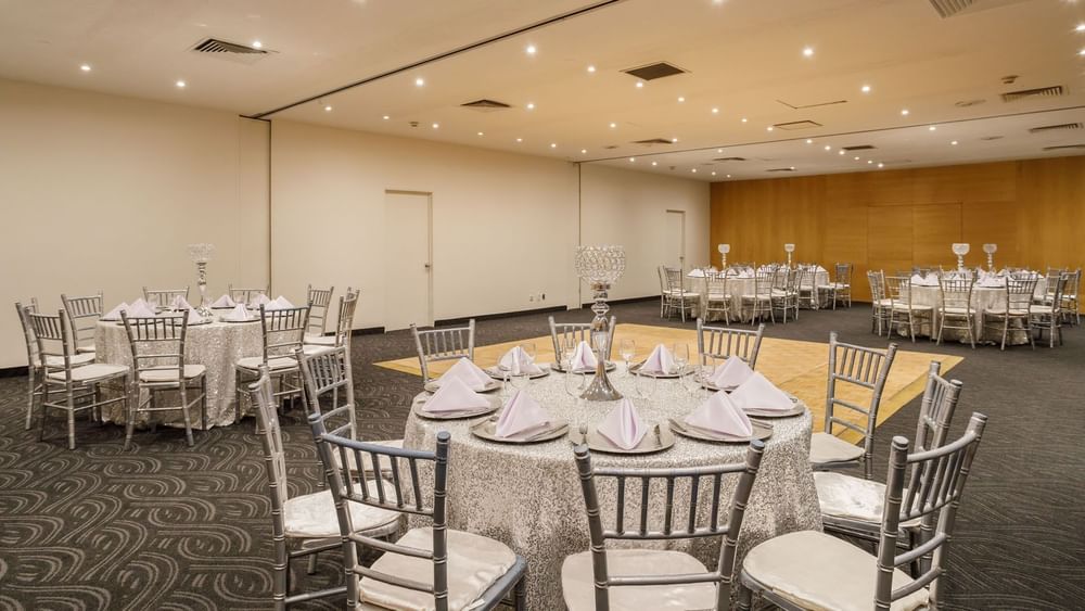 Spacious Nueva York event room featuring round tables with silver linens and a wooden dance floor at Real Inn Torreon