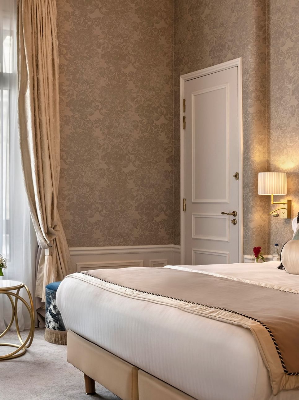 Elegant hotel bedroom with upholstered bed, decorative pillows, gold-toned curtains, and soft lighting creating a refined ambiance.