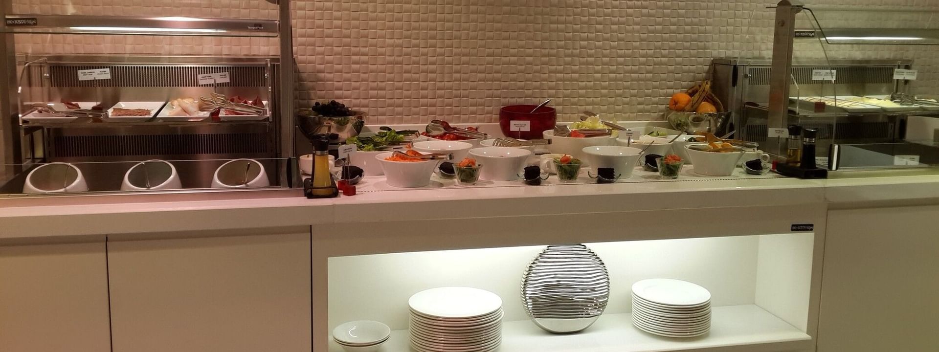 Salad bowls by stacks of white plates under a glowing display shelf in Breakfast & In-Room Dining at Warwick Ankara