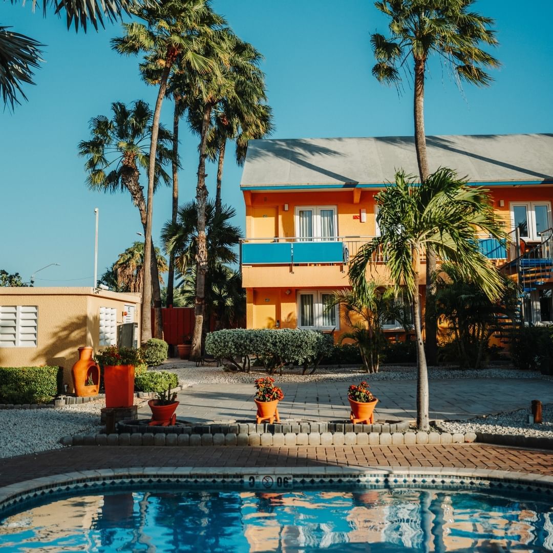 Yellow hotel with blue accents, palm trees, and a pool under a clear blue sky.
