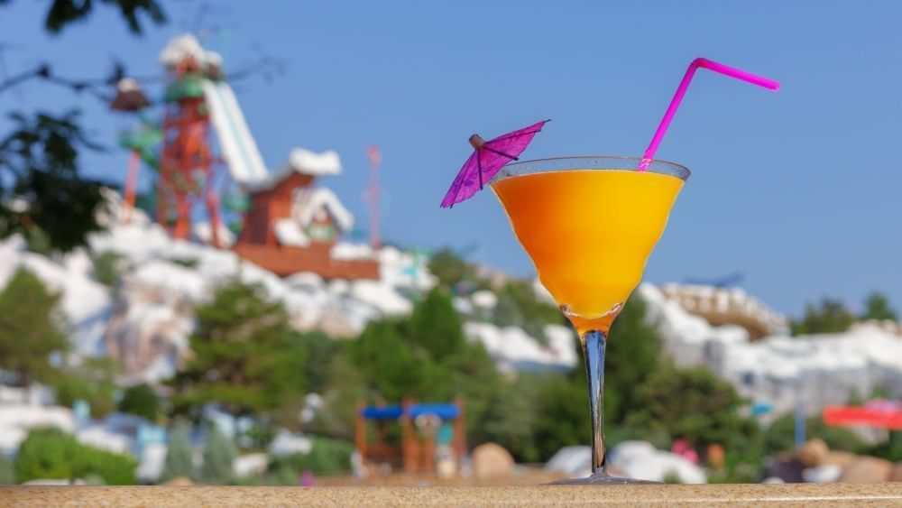 An orange cocktail in a martini glass with a pink umbrella and straw sits in the foreground with a large water slide ski ramp out of focus in the background.