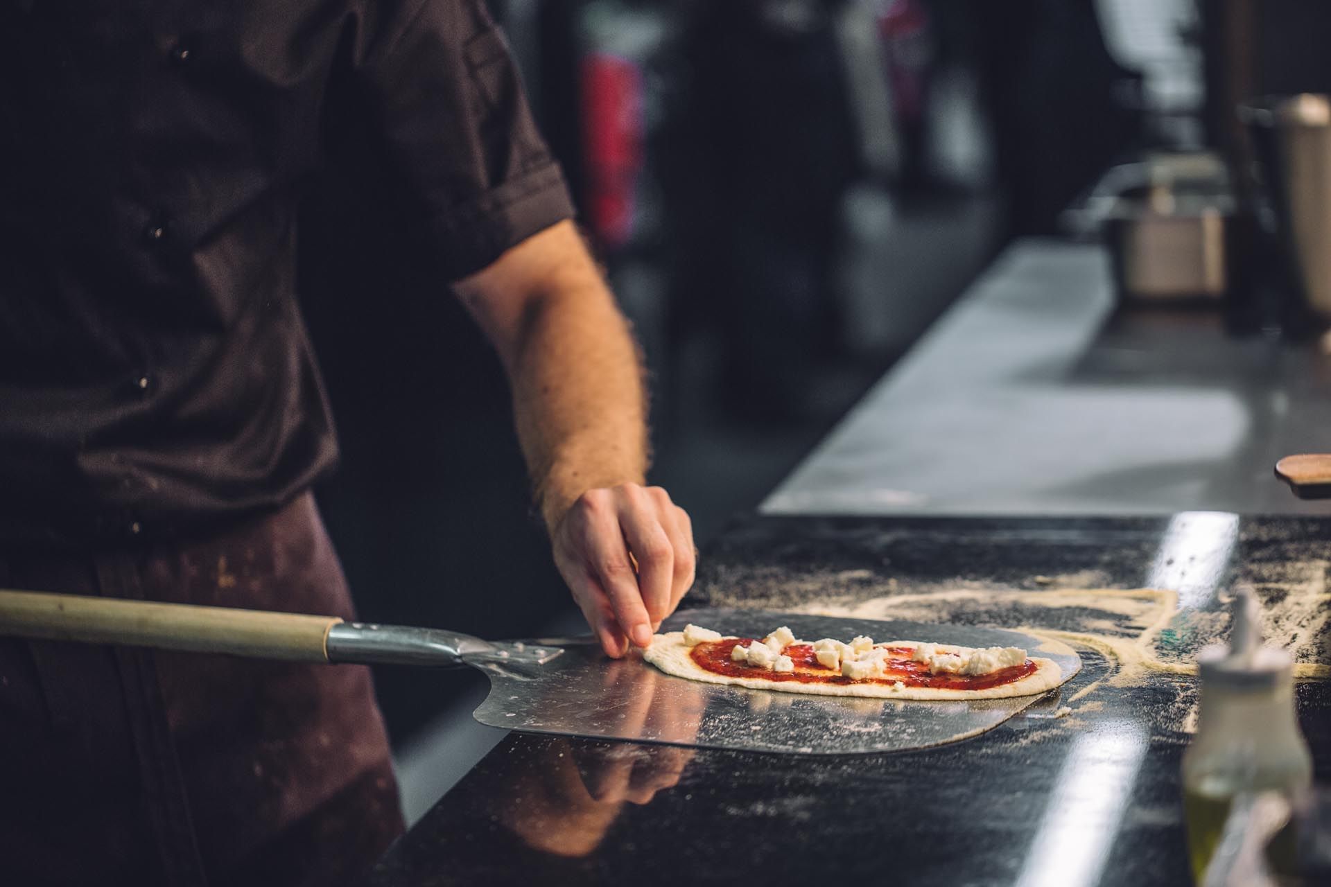Chef spreads tomato sauce & cheese on pizza dough using a pizza peel with flour on the countertop at Monmouth Kitchen