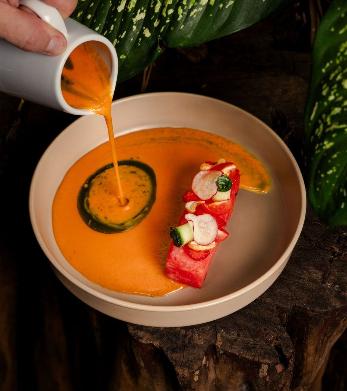 Hand pours an orange-hued gazpacho into a beige bowl containing a piece of watermelon served at Cala Luna Boutique Hotel