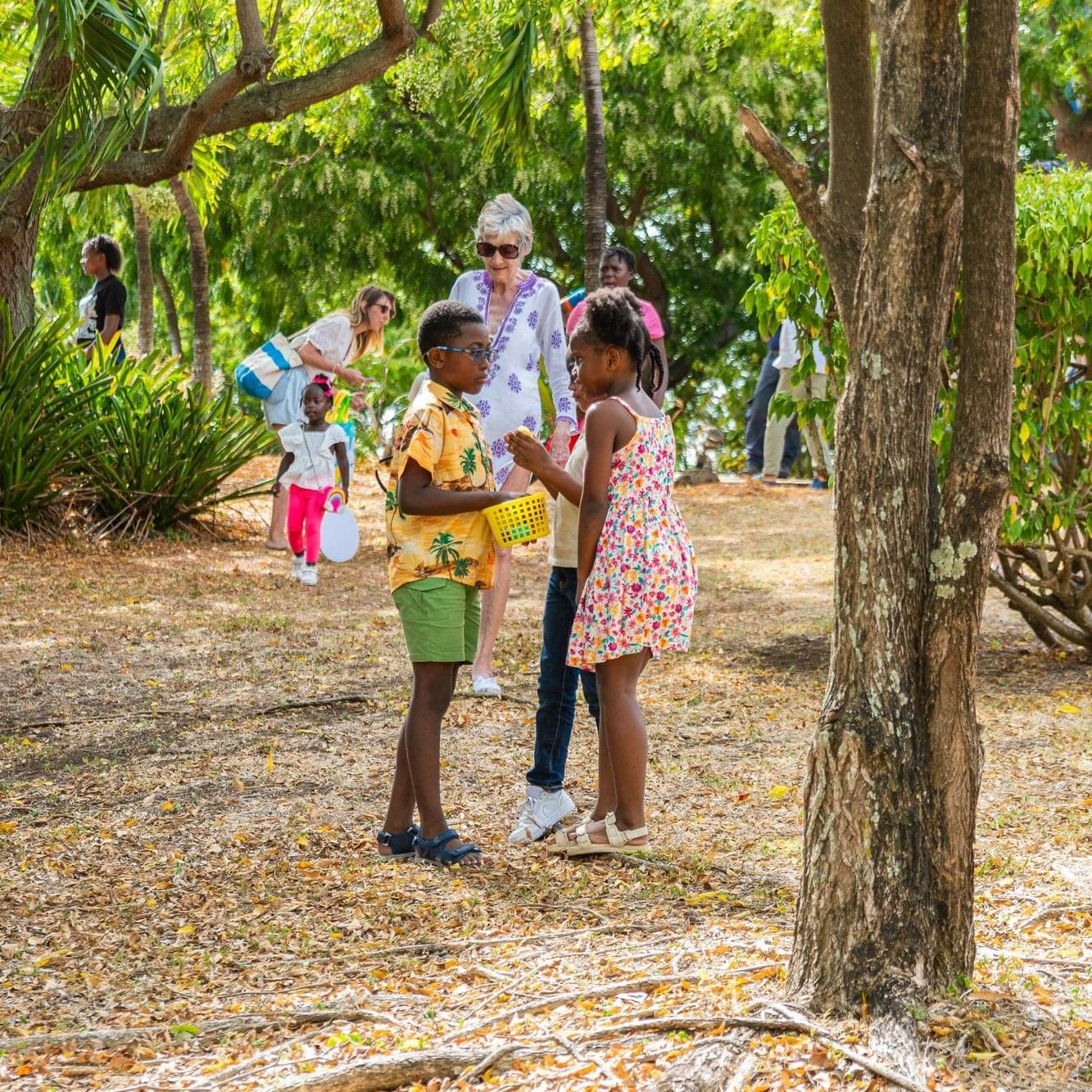 Children and adults enjoying outdoor activities together in a lush tropical garden setting with tall trees and foliage.