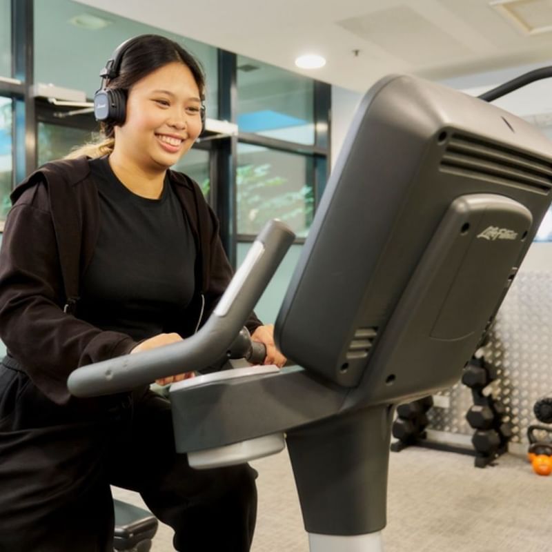 Smiling student using exercise bike in modern fitness center with large windows and workout equipment.
