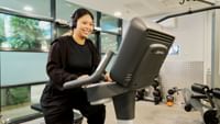 Smiling student using exercise bike in modern fitness center with large windows and workout equipment.