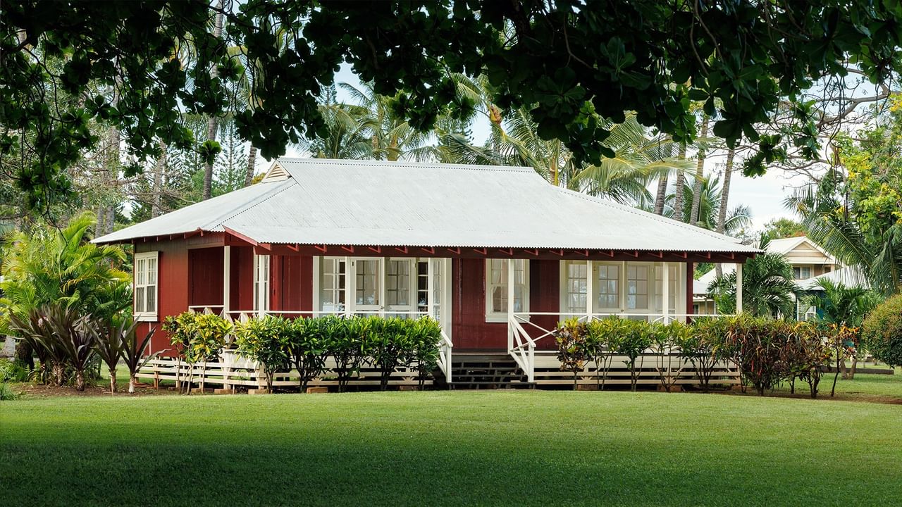 Red plantation-style cottage with white trim and wraparound porch surrounded by lush tropical gardens and green lawn.