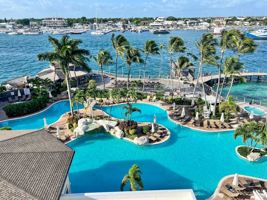 View of Warwick Paradise Island Bahamas, with a large pool by white umbrellas under palm trees surrounding a tropical harbor