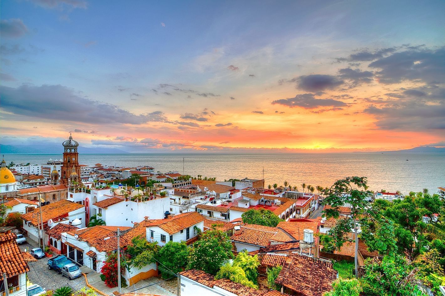 Vista panorámica al atardecer de Puerto Vallarta con techos de teja roja, la Iglesia de Nuestra Señora de Guadalupe y el Océano Pacífico brillando bajo un cielo colorido.