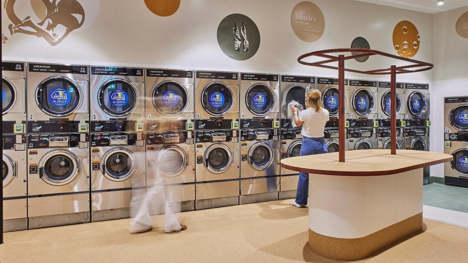Woman standing behind a counter in a laundromat, while another woman walks by in a blur.