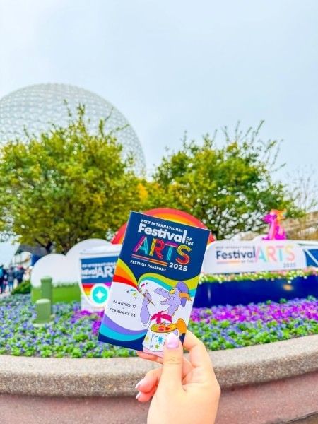 A hand holding a promotional item for the EPCOT Festival of the Arts in front of the EPCOT ball at Walt Disney World.