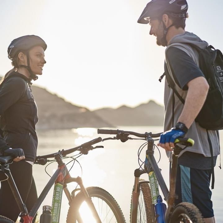 Coppia con biciclette da montagna vicino a un lago al tramonto, entrambi indossano caschi.