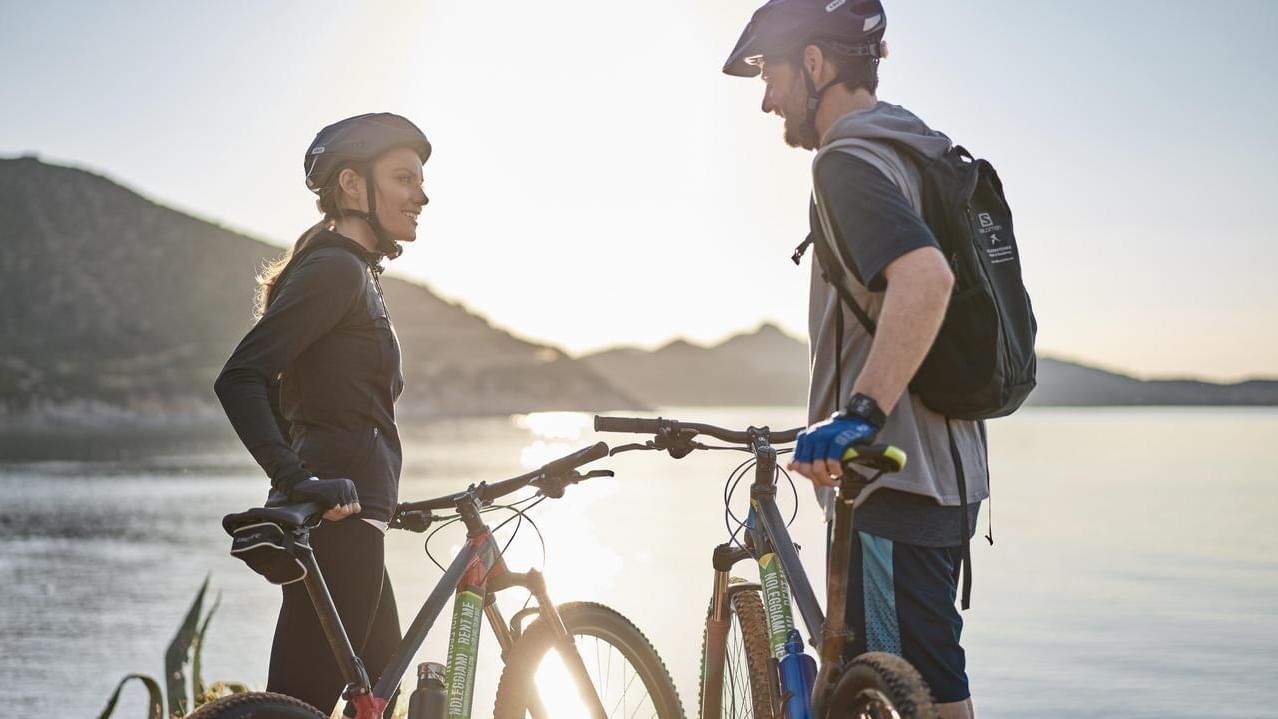Coppia con biciclette da montagna vicino a un lago al tramonto, entrambi indossano caschi.