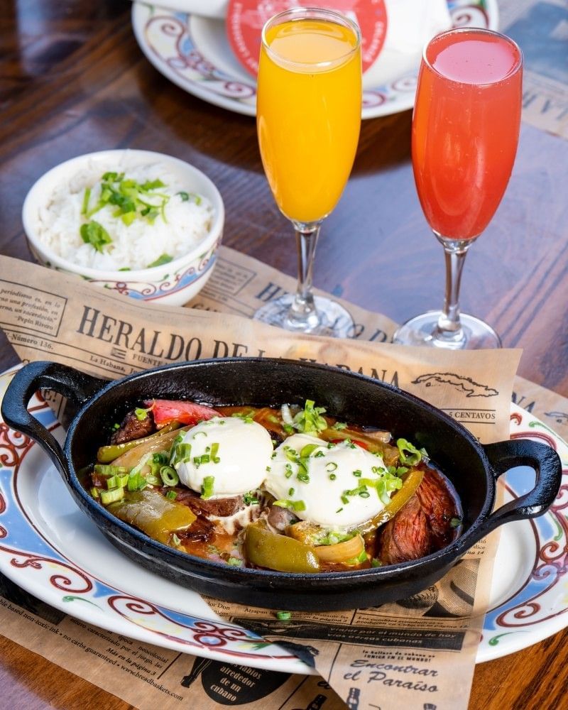 A skillet with eggs and vegetables sits on a table beside an orange mimosa and a bowl of rice.