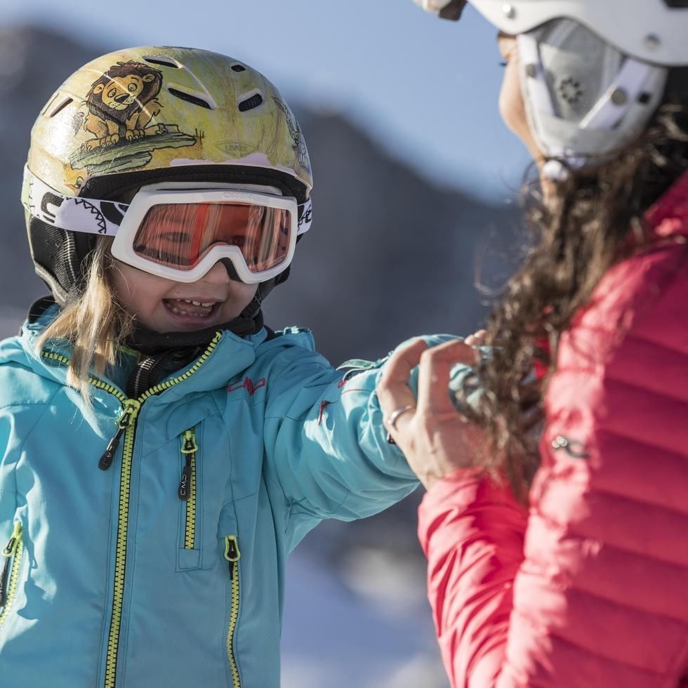 Lächelndes Kind mit Skihelm und Skibrille hält Hand eines Erwachsenen auf der Skipiste an einem sonnigen Wintertag.