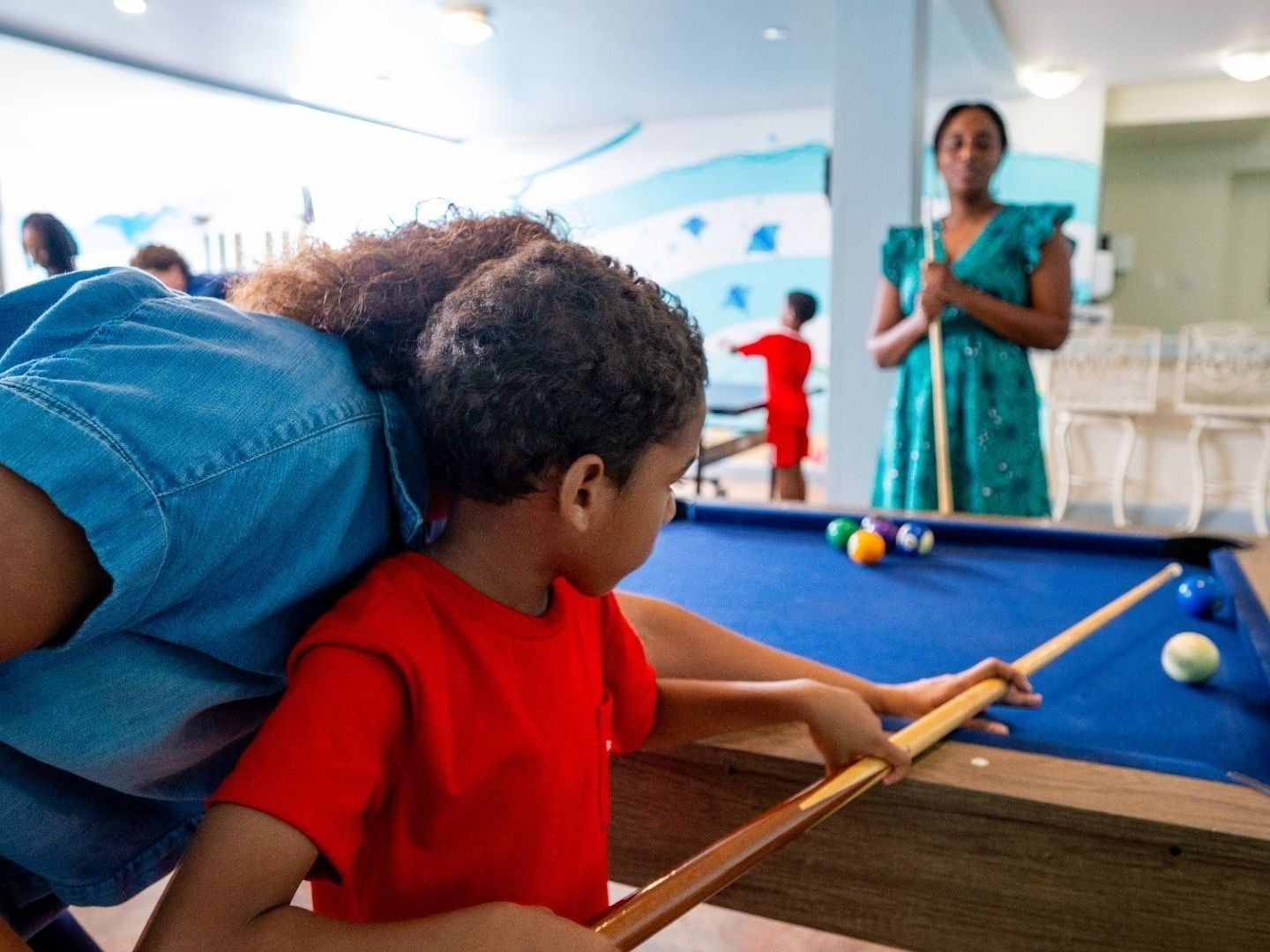 A child with a woman playing pool with other kids in the background.