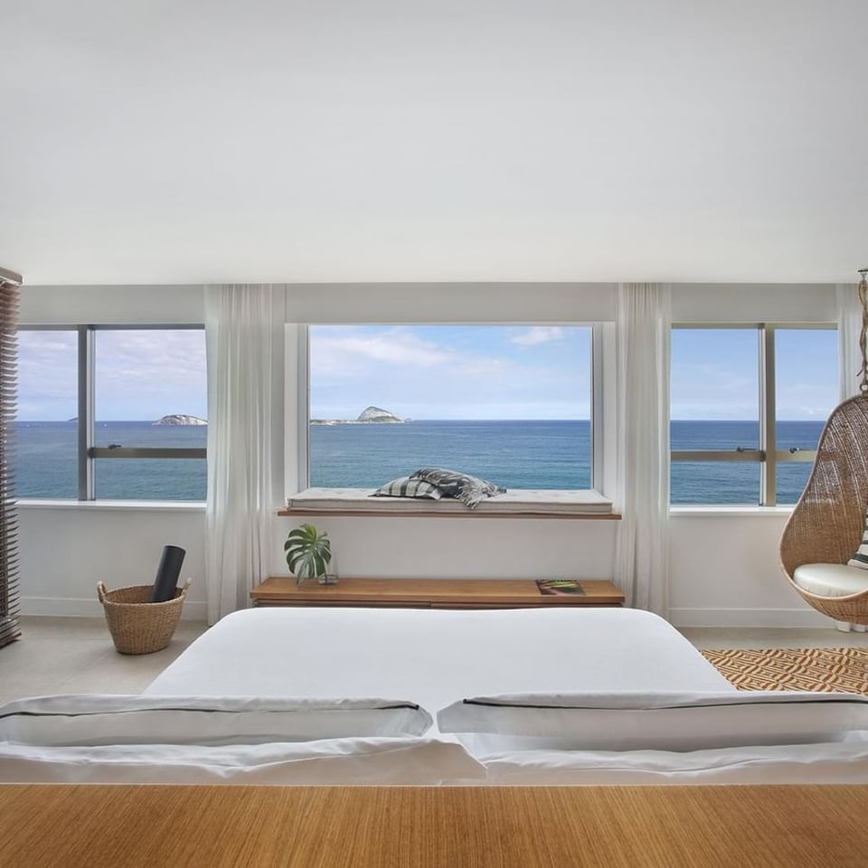 Suíte master luxo do Janeiro Hotel com cama king size e vista para o mar com uma cadeira suspensa ao lado 