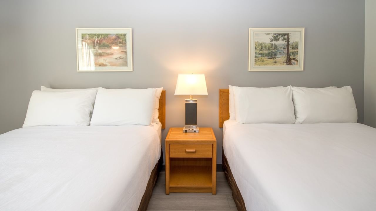 Two beds with white linens and pillows, a nightstand with a lamp, and two framed pictures on the wall.
