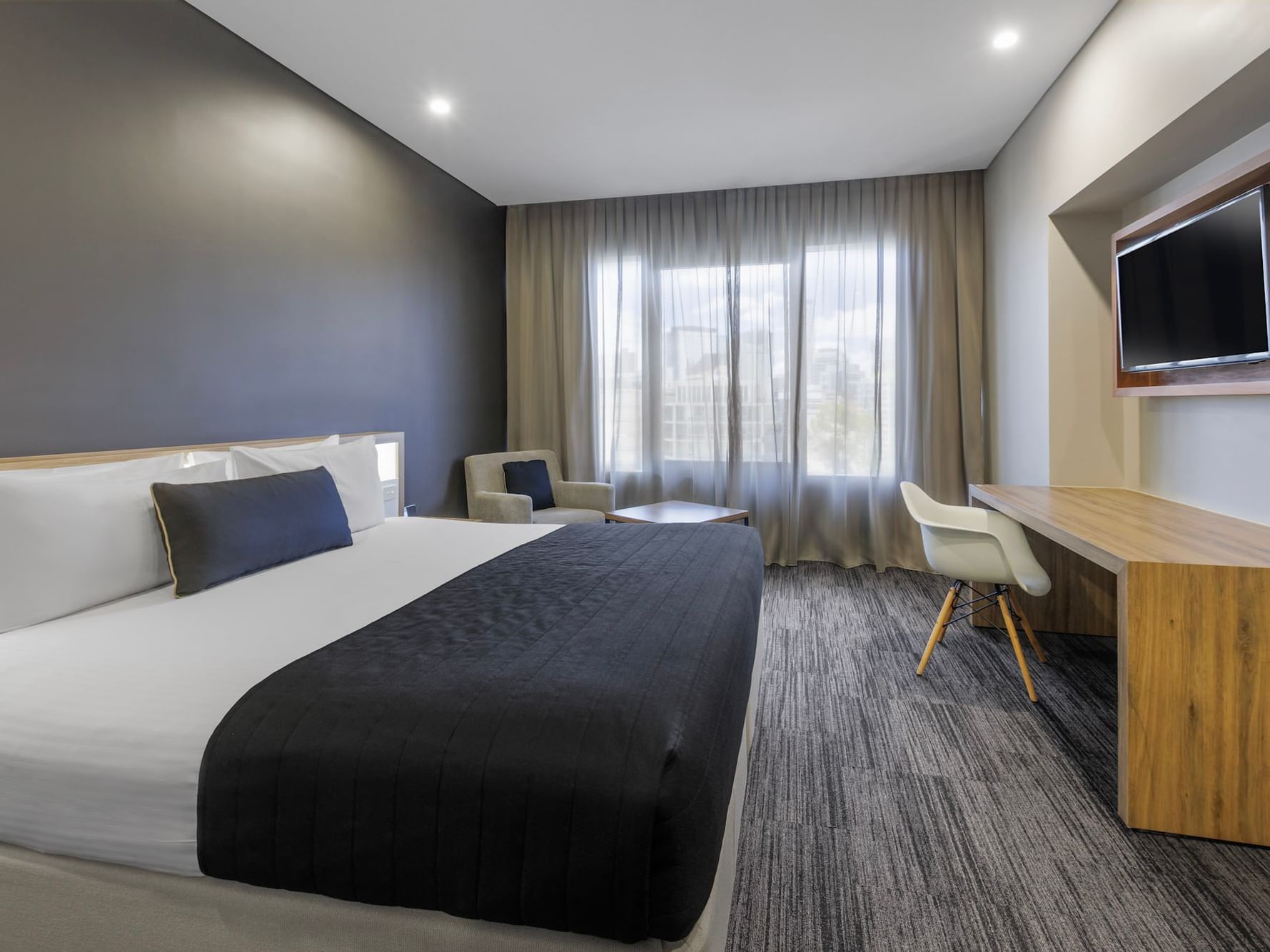 Modern Executive King Room at Hotel Grand Chancellor Brisbane featuring a black bed runner, charcoal accent wall, integrated wooden desk, and large window with sheer curtains.