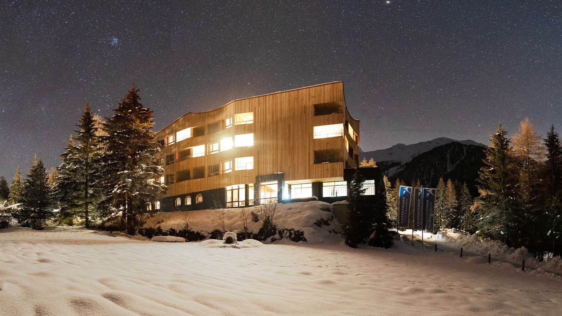 A wooden building with lit windows and snow-covered ground at night under a starry sky.