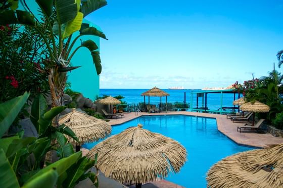 Outdoor swimming pool surrounded by tropical thatched umbrellas and green palm trees at the Emerald Beach Resort