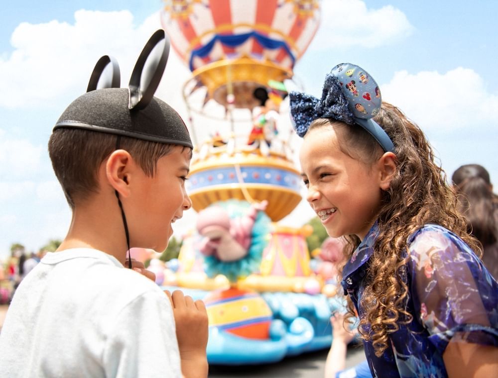 A boy and girl in Minnie and Mickey ears smile at each other in front of an out-of-focus parade float.