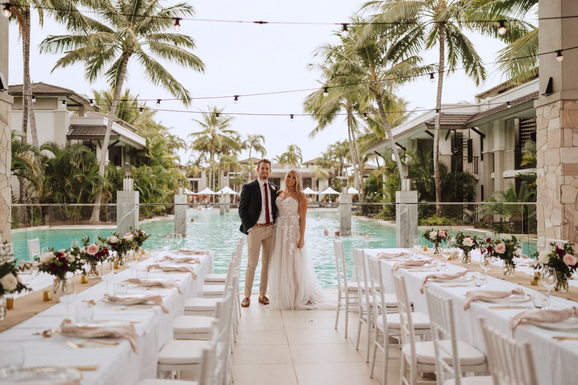 Sophie and James stands hand in hand by poolside with tables & chairs in Boardwalk at Pullman Port Douglas Sea Temple Resort & Spa