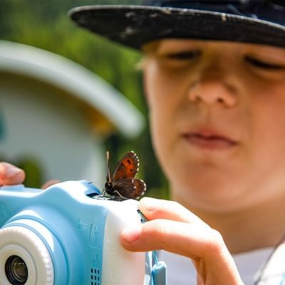 A boy holds a butterfly on a camera lens with his hand in an outdoor setting.