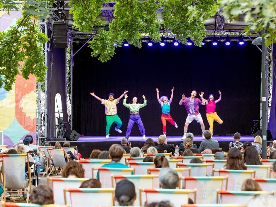 Dance group performing on an open-air stage at Kultursommer Wien, with audience seated in colorful deck chairs.