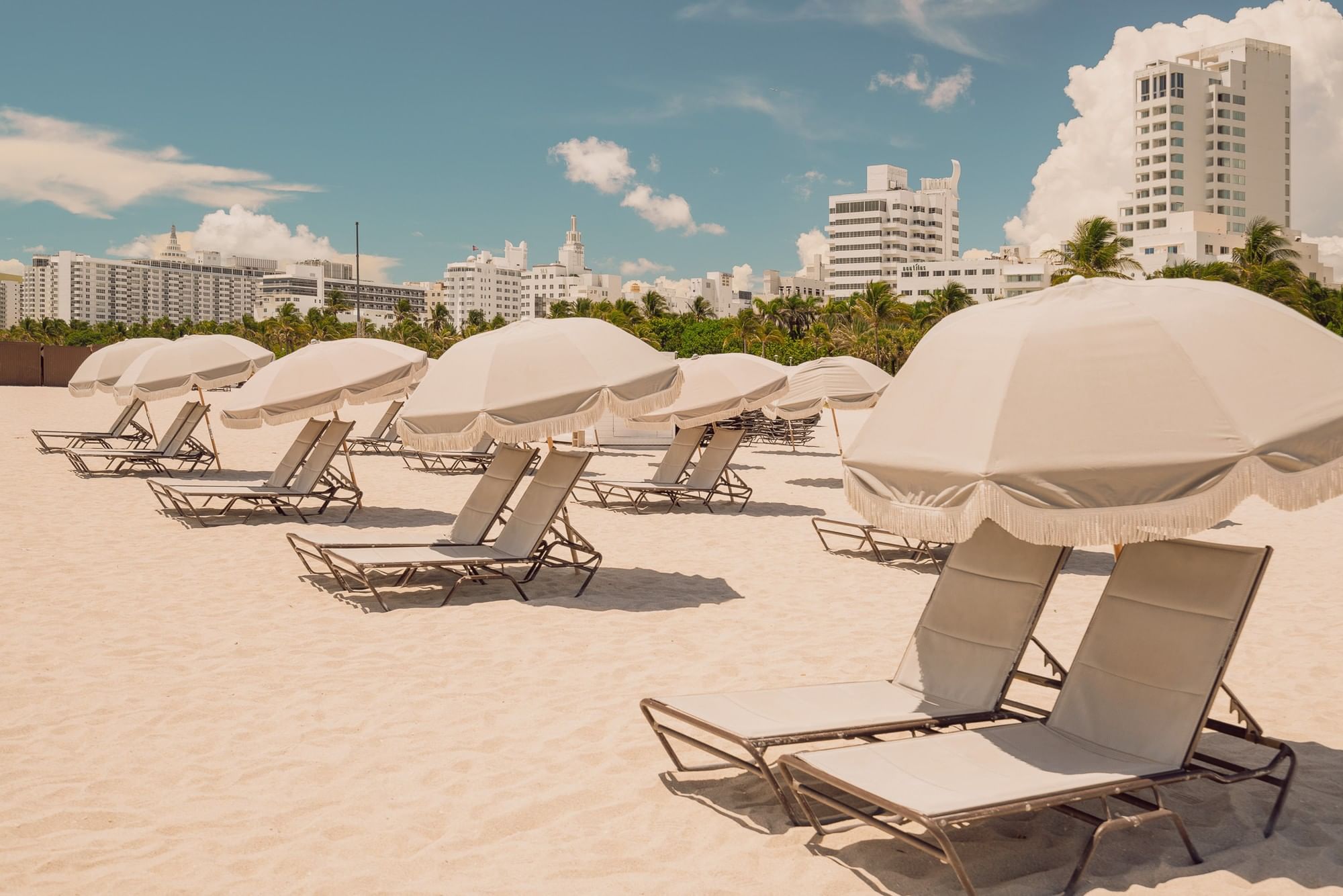 A beach scene with white sand, lounge chairs, and umbrellas, with a city skyline in the background.