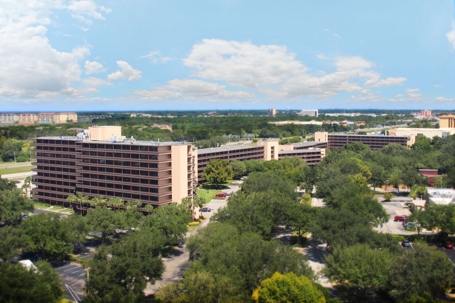 A wide aerial view of a multi-story hotel building with a parking lot and trees.