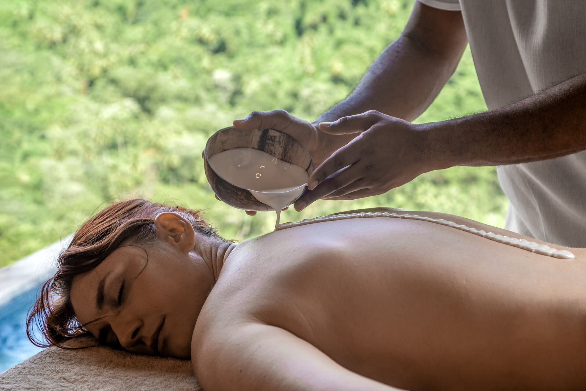 A woman is receiving a coconut milk massage by a therapist near a pool.