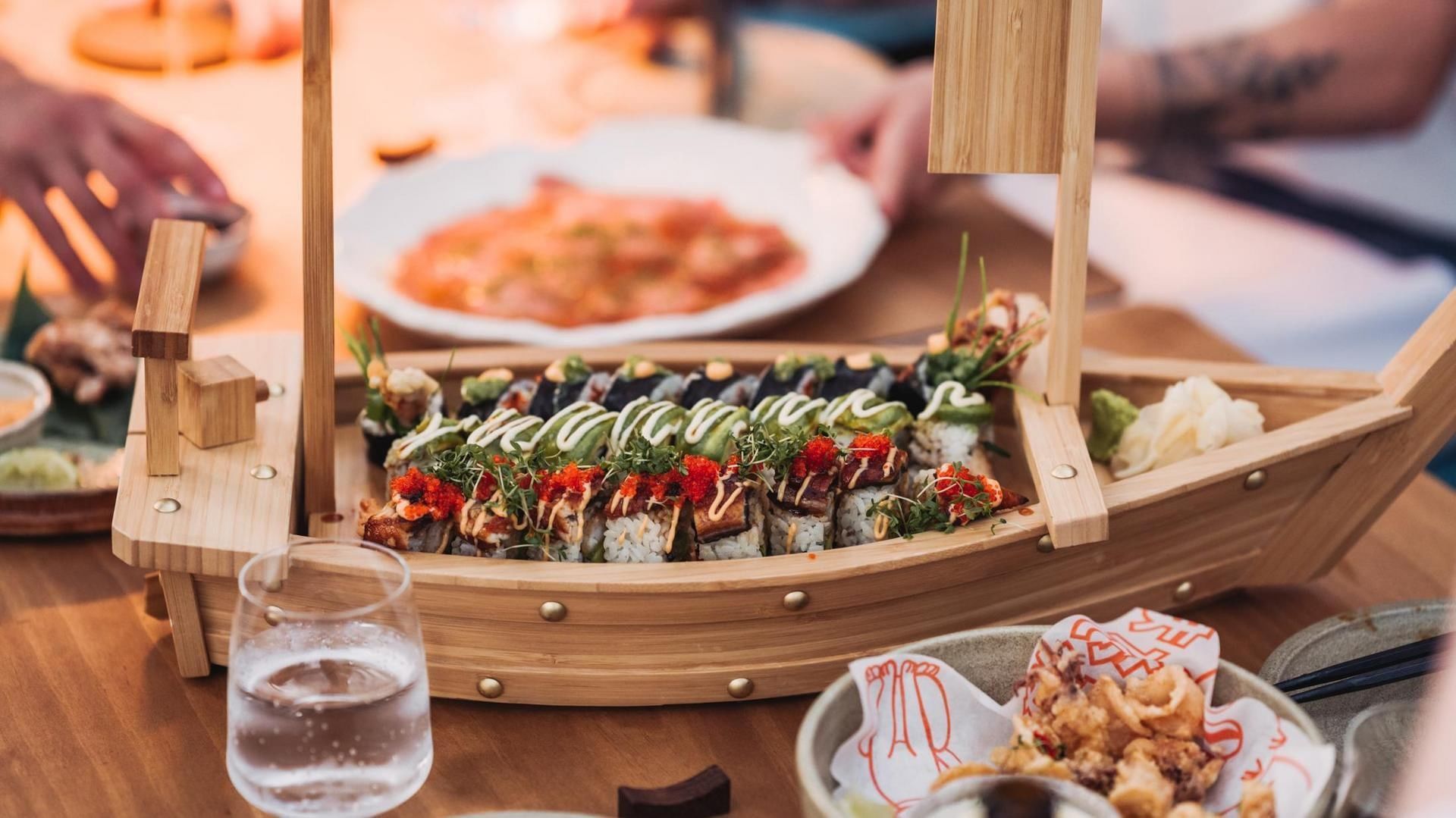 Barchetta di legno con sushi e sashimi su un tavolo da pranzo con altri piatti e bicchiere d'acqua.