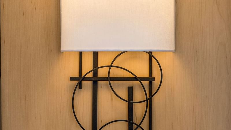 White cylindrical lampshade on black metal frame with abstract circular design, mounted on wooden wall.