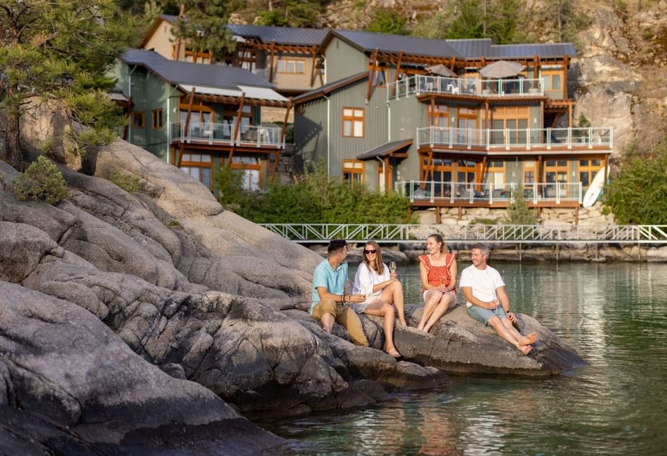 Four people sitting on rocks near a lake, with a resort in the background.