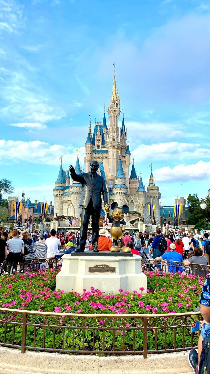The image shows a statue of Walt Disney and Mickey Mouse in front of Cinderella Castle at Magic Kingdom, Disney World. People are visible in the background. Pink flowers are in the foreground.
