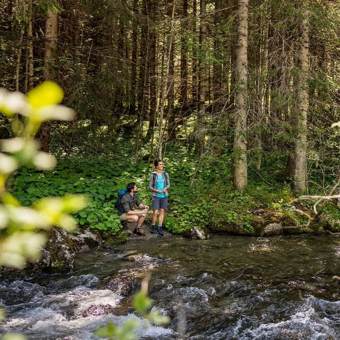 Two hikers by a river in a forest, one seated and the other standing.