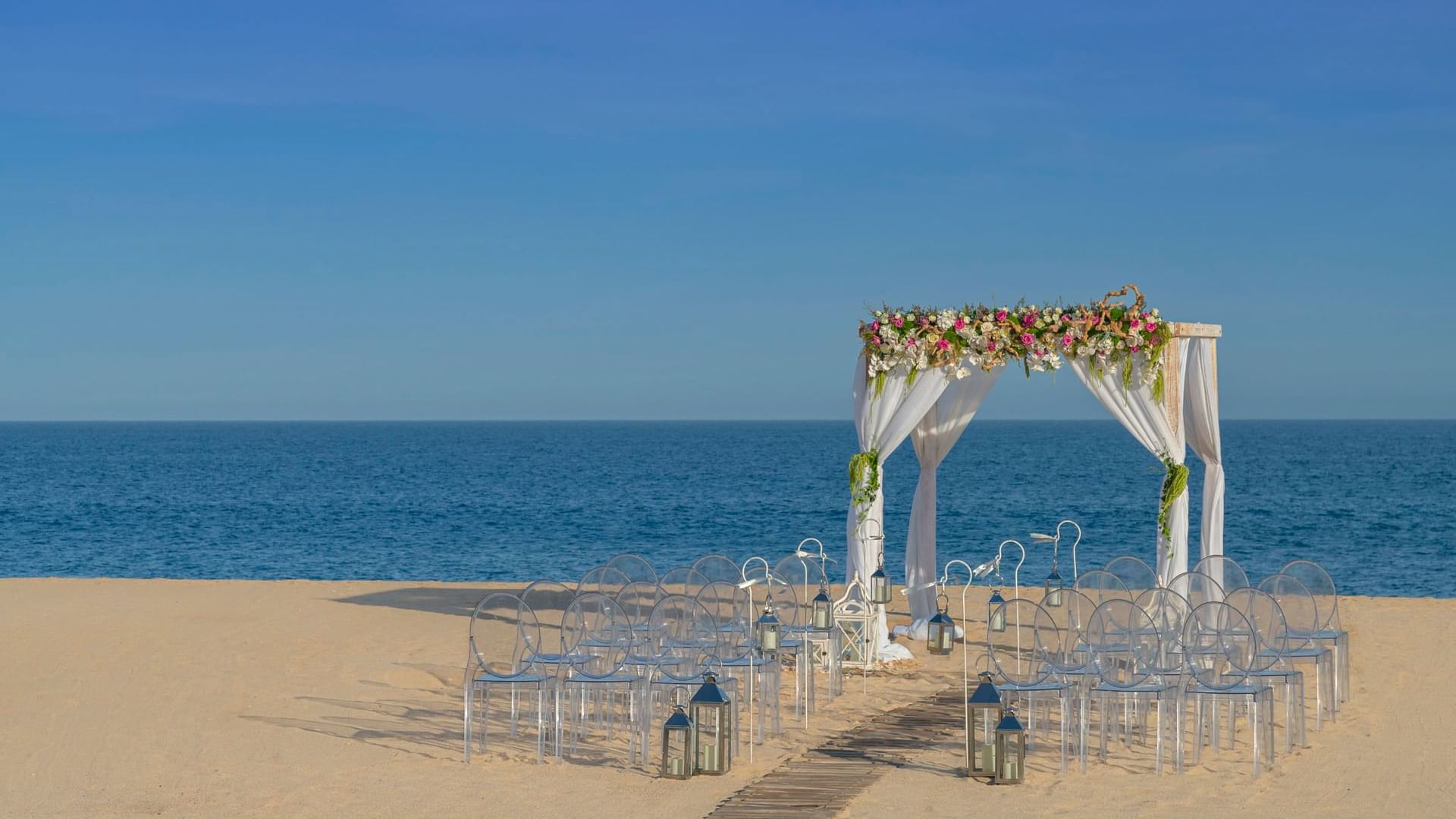 A romantic beach ceremony setup at Hacienda del Mar Los Cabos with a floral arch and clear chairs