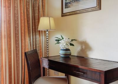 Wooden desk with a chair, featuring a vase of white flowers & a stylish lamp, creating a cozy workspace at Bay Gardens Hotel