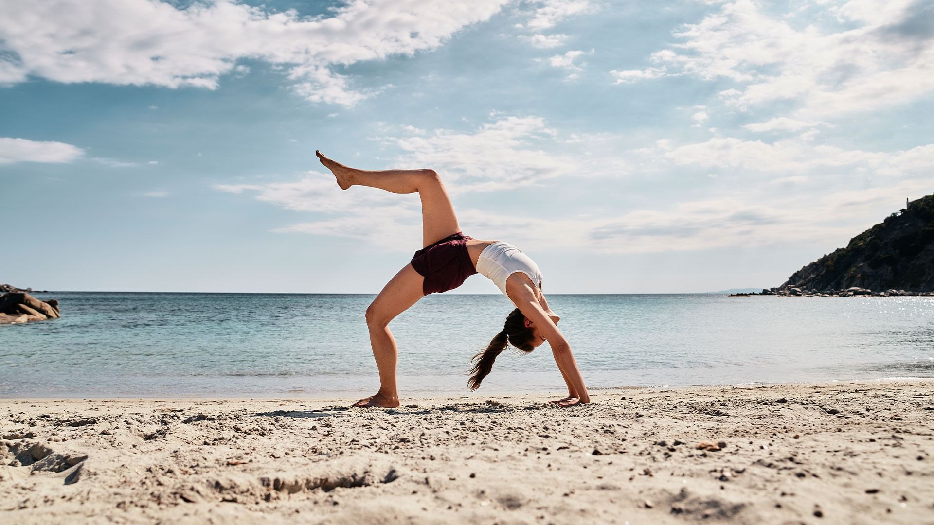 Woman doing a handstand on sandy beach with ocean and mountain in background.