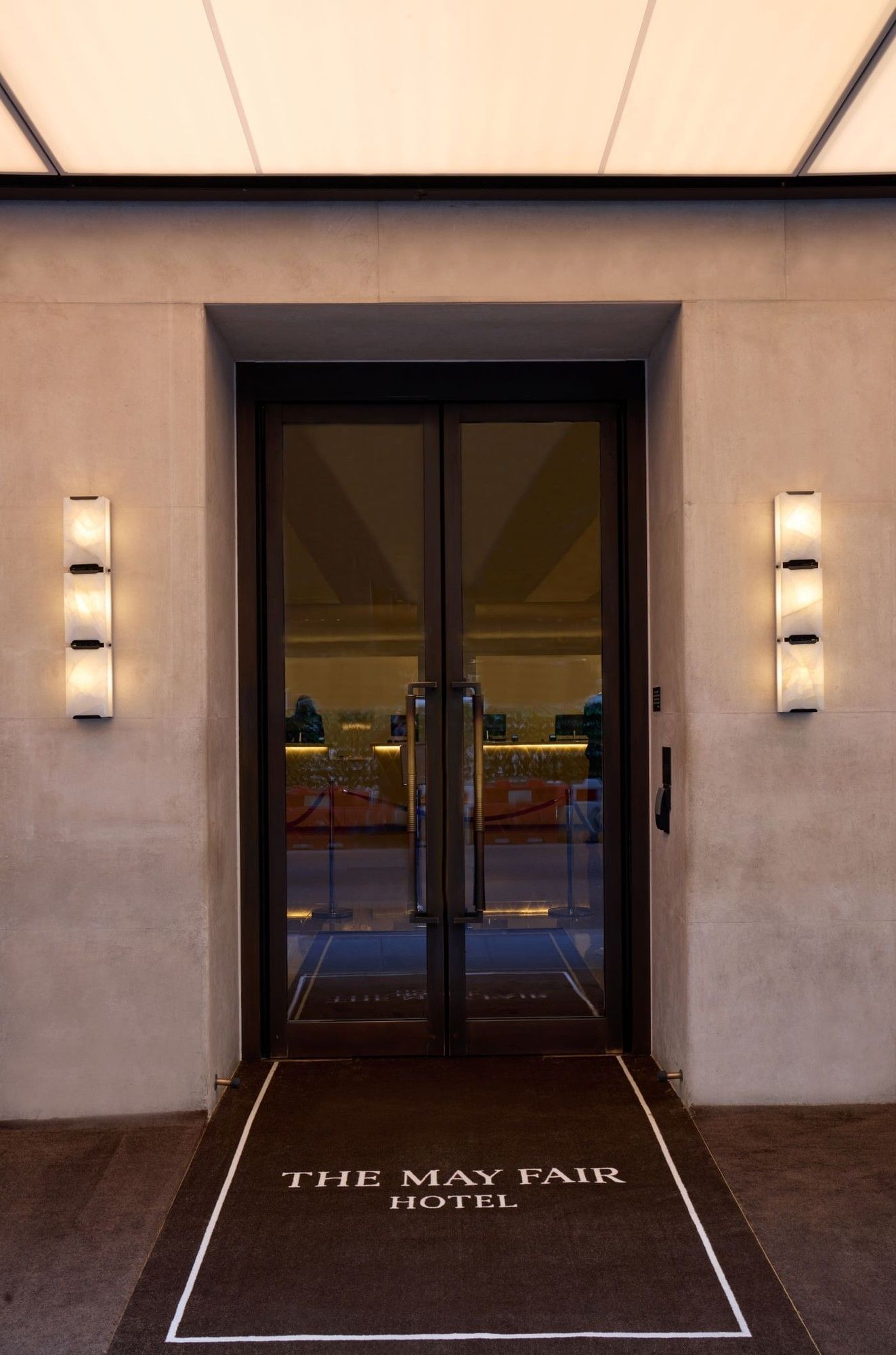 Entrance of The May Fair Hotel with glass doors and lit wall sconces.