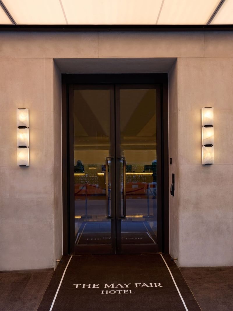 Entrance of The May Fair Hotel with glass doors and lit wall sconces.