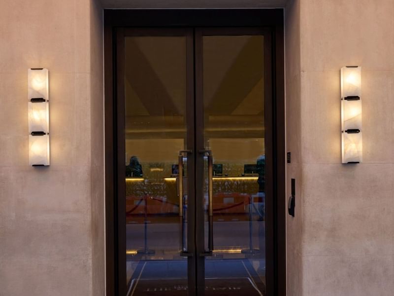 Entrance of The May Fair Hotel with glass doors and lit wall sconces.
