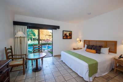 Spacious hotel room featuring a king-size bed, warm lighting, and a small wooden dining table with chairs.